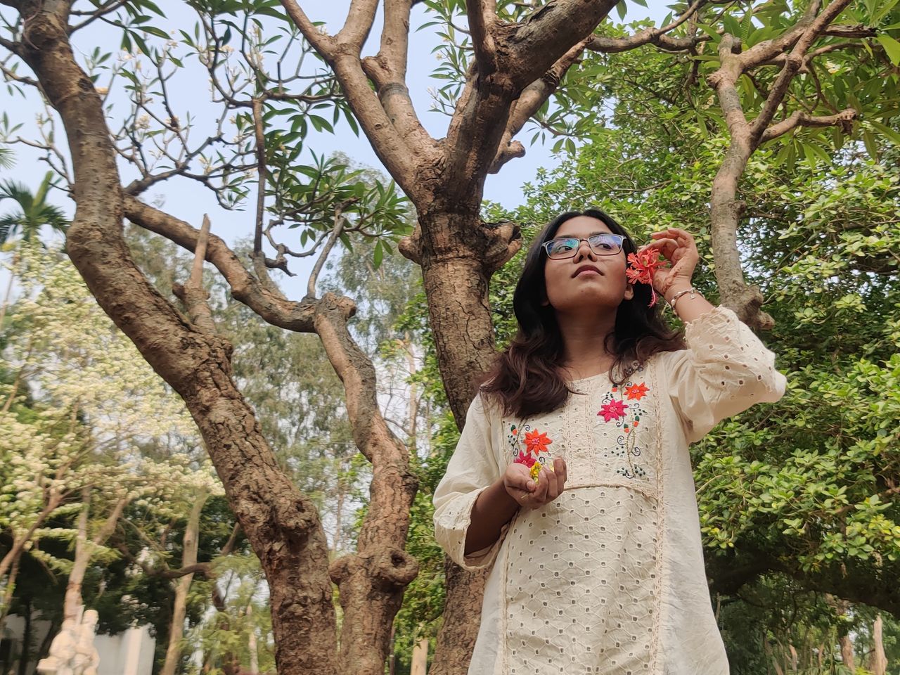 A young woman in a light dress holding flowers while standing under a tree on a sunny day.